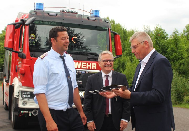 Das Bild zeigt drei Männer bei der Übergabe eines Schlüssels vor einem großen Feuerwehrfahrzeug.