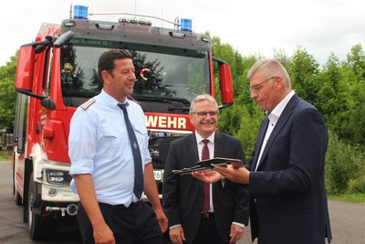 Das Bild zeigt drei Männer bei der Übergabe eines Schlüssels vor einem großen Feuerwehrfahrzeug.