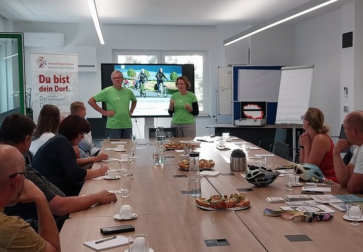 Eine Gruppe von Menschen sitzt um eine große Tafel herum. Am Kopfende halten zwei Menschen in grünen T-Shirts einen Vortrag. Im Hintergrund ist ein großer Bildschirm mit einer Präsentation zu sehen. 