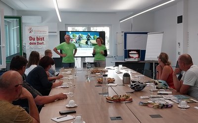Eine Gruppe von Menschen sitzt um eine große Tafel herum. Am Kopfende halten zwei Menschen in grünen T-Shirts einen Vortrag. Im Hintergrund ist ein großer Bildschirm mit einer Präsentation zu sehen.