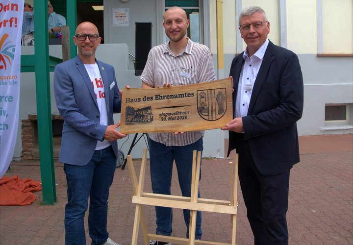 Drei Männer halten eine Holztafel mit der Aufschrift "Haus des Ehrenamtes" in der Hand. 
