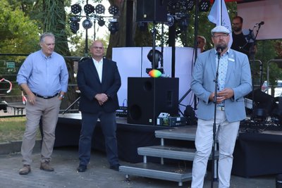Drei Männer mit Hemd und Jeacket stehen vor einer Bühne. Einer der Herren spricht in ein Mikrofon.