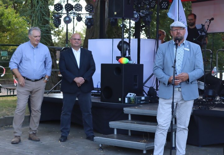 Drei Männer mit Hemd und Jeacket stehen vor einer Bühne. Einer der Herren spricht in ein Mikrofon.