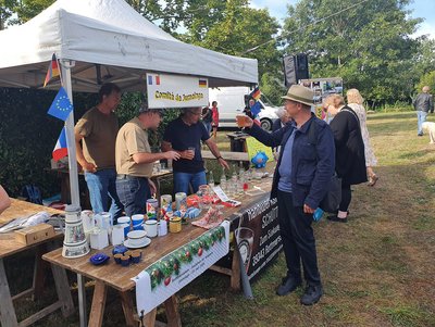 Drei Männer bieten an einem Stand Flohmarkt-Artikel an. Vor Ihnen steht ein Mann mit Hut, der ihnen mit einem Becher Bier zuprostet.