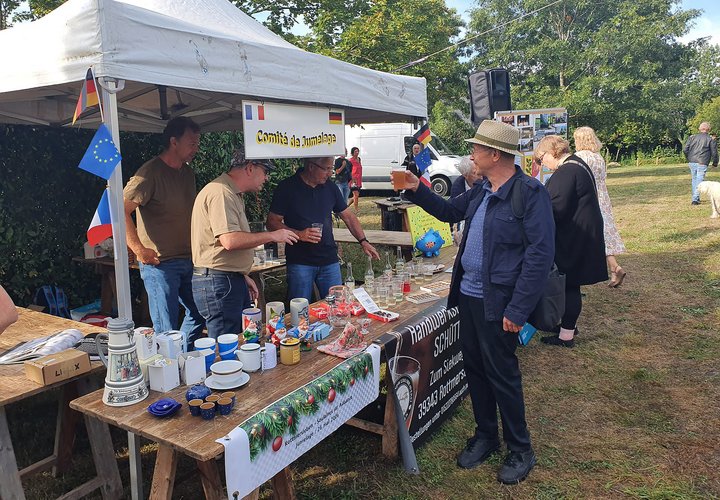 Drei Männer bieten an einem Stand Flohmarkt-Artikel an. Vor Ihnen steht ein Mann mit Hut, der ihnen mit einem Becher Bier zuprostet.