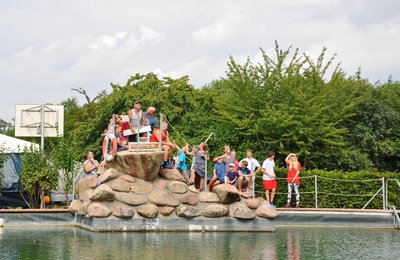 Am Rand eines Schwimmbeckens haben sich Kinder und ein Erwachsener um einen Sprungturm versammelt. 