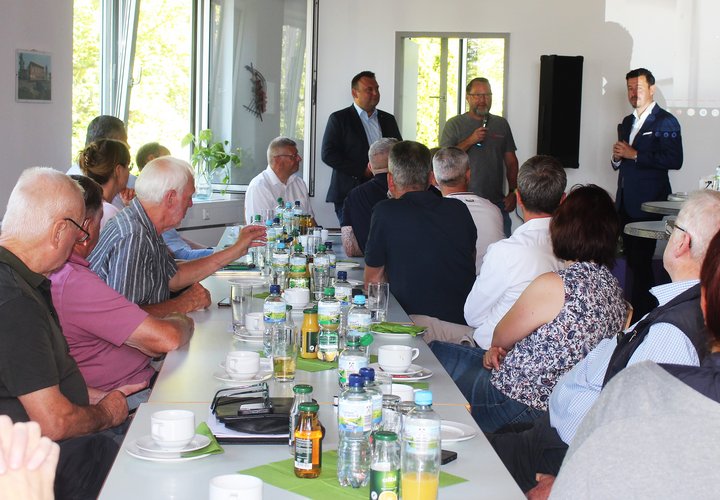 Das Foto zeigt im Hintergrund drei Männer, die vor mehreren Menschen an einer Kaffeetafel referieren.