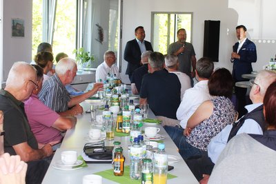 Das Foto zeigt im Hintergrund drei Männer, die vor mehreren Menschen an einer Kaffeetafel referieren.