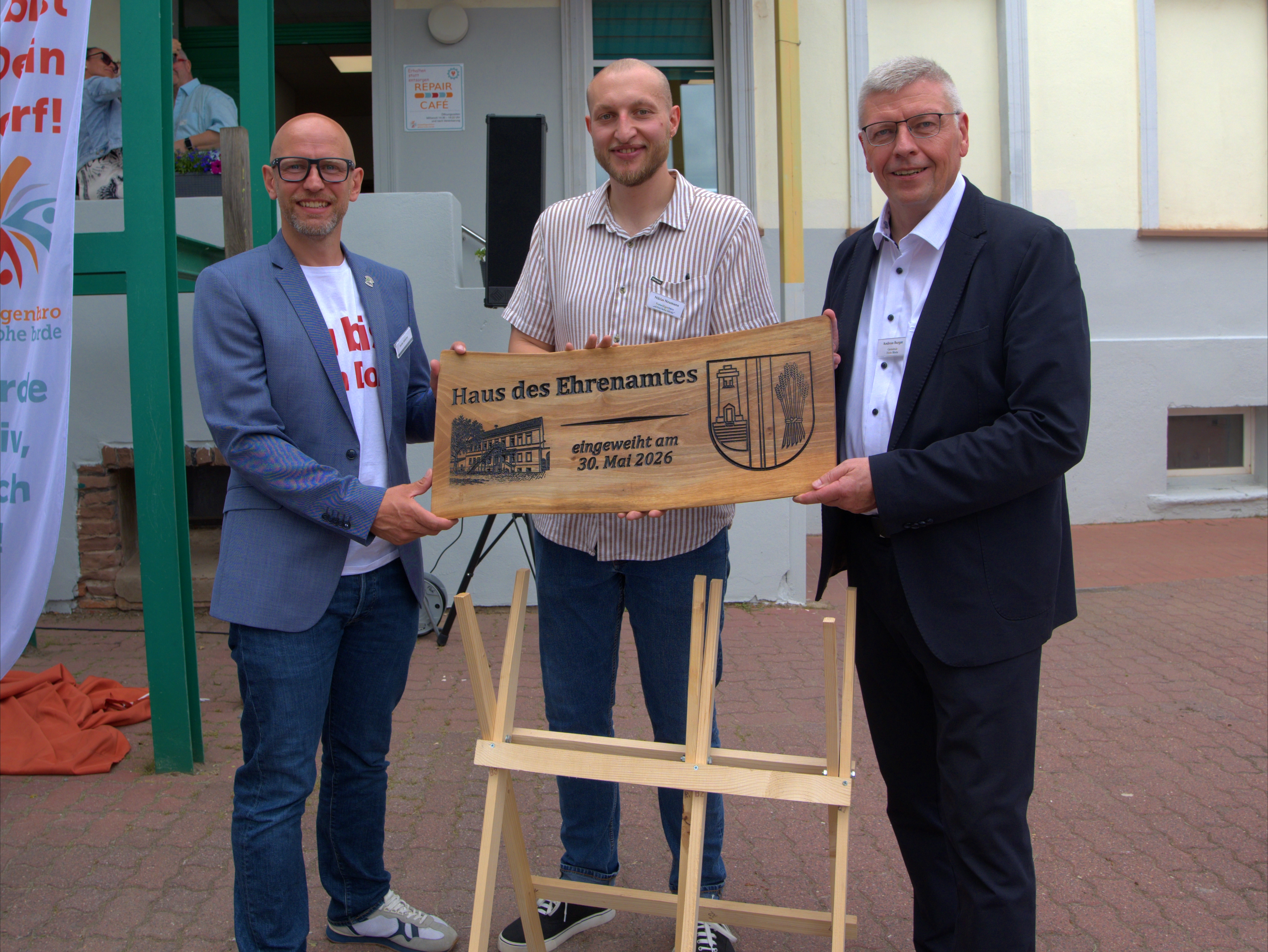 Drei Männer halten eine Holztafel mit der Aufschrift "Haus des Ehrenamtes" in der Hand.